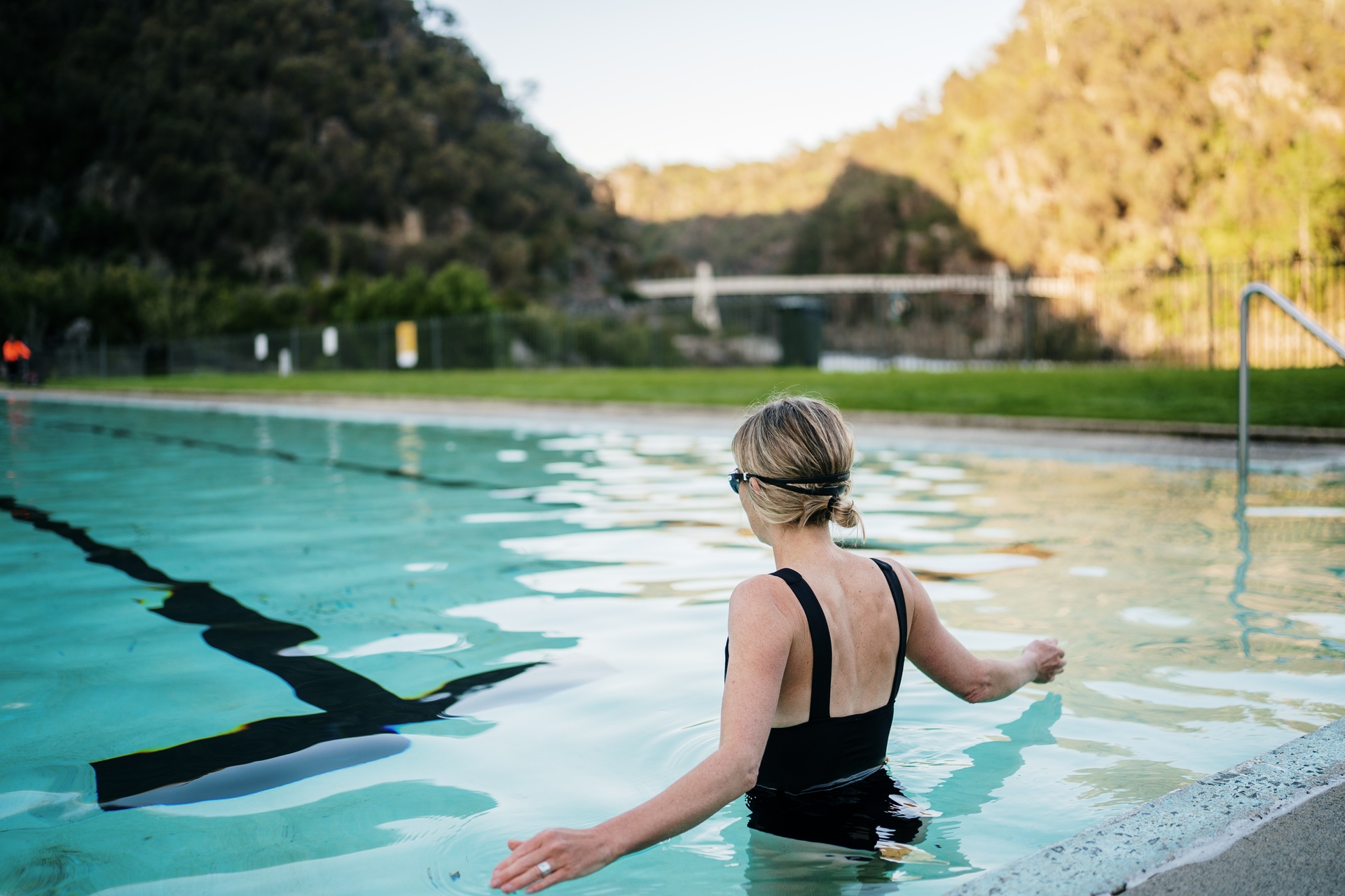 A female swimmer standing in the pool at the Launceston Gorge. She has blonde hair tied in a bun and her arms open just touching the water. The Suspension Bridge can be seen in the distance.